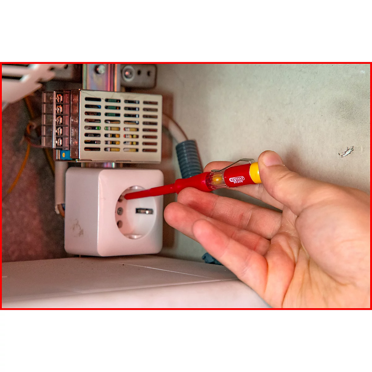 Hand mit rotem Schraubendreher testet Steckdose. Über der Steckdose ein Schaltkasten.